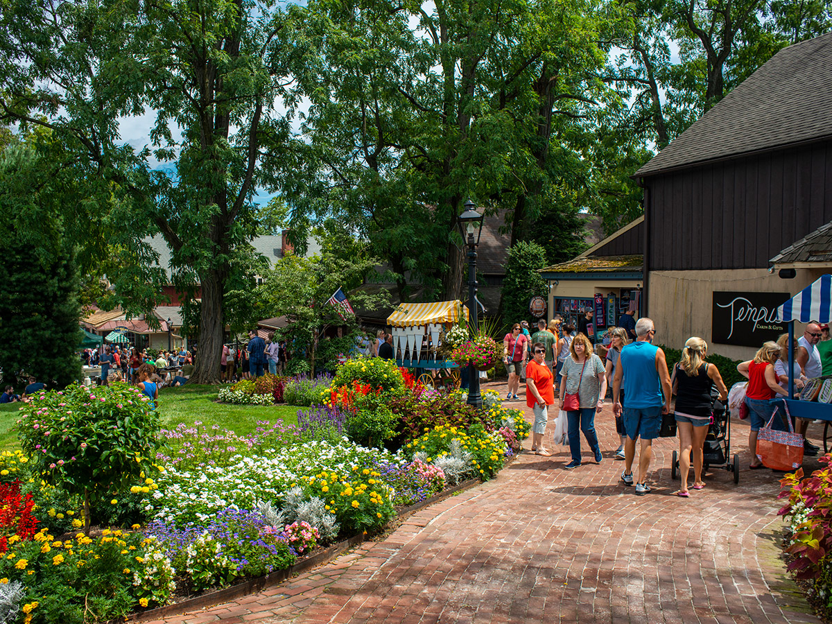 People walk along a brick walkway through a grassy lawn and shrubs to a colonial style buildings filled with shops at Peddler's Village.