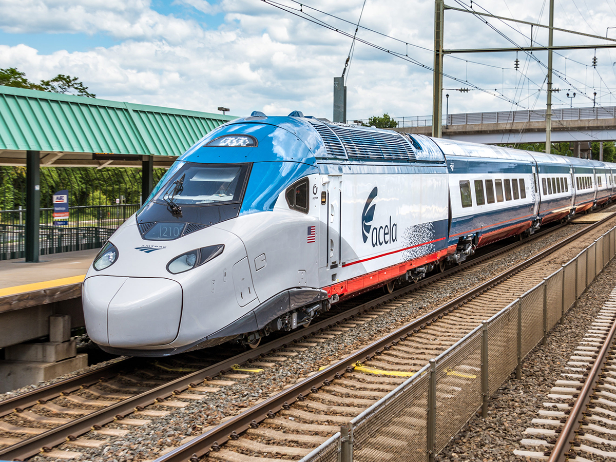 A blue, white and red Acela train stops at a train station along the train tracks.