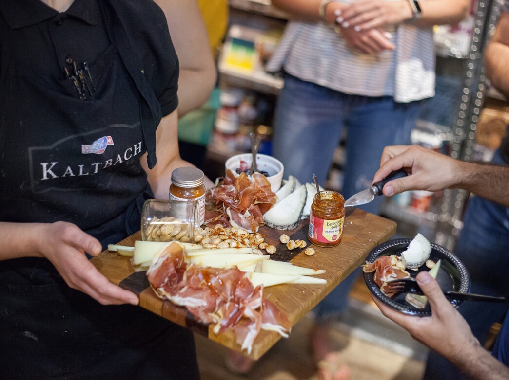 A person wearing a black apron holds out a charcuterie board with meat, cheese, nuts and jam while another person selects items from the board.