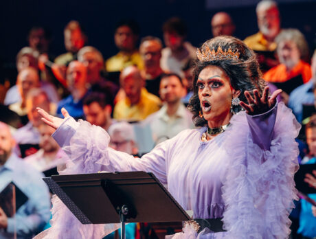 Drag queen Cookie DiOrio sings jubilantly in front of a chorus of men. The queen wears a crown and a frilled purple dress