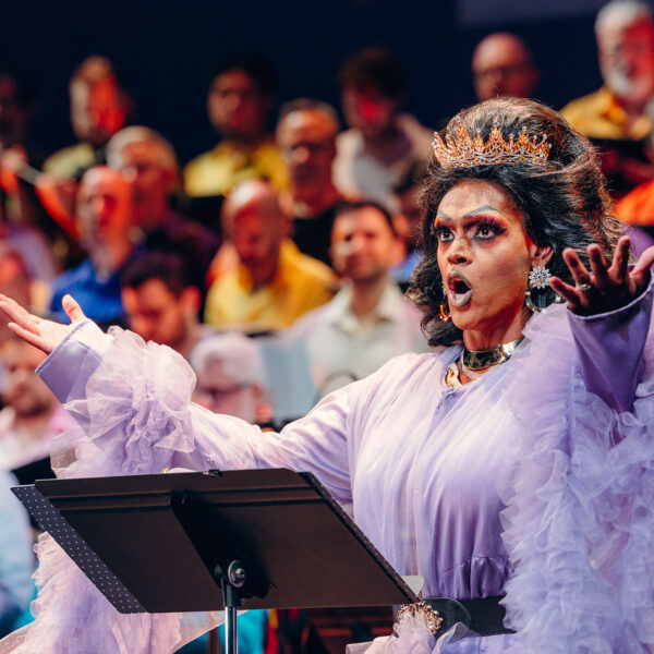 Drag queen Cookie DiOrio sings jubilantly in front of a chorus of men. The queen wears a crown and a frilled purple dress