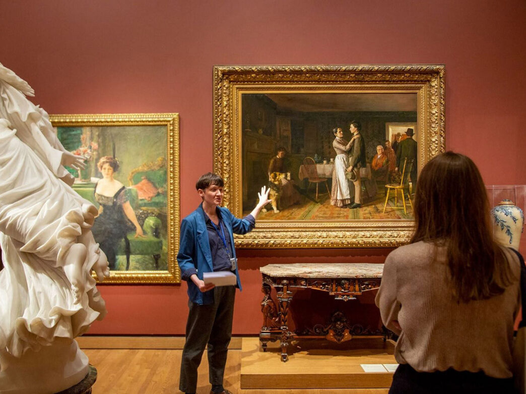 A museum guide points to an old painting of people standing in a historic house. A visitor observes the guide and gallery from afar