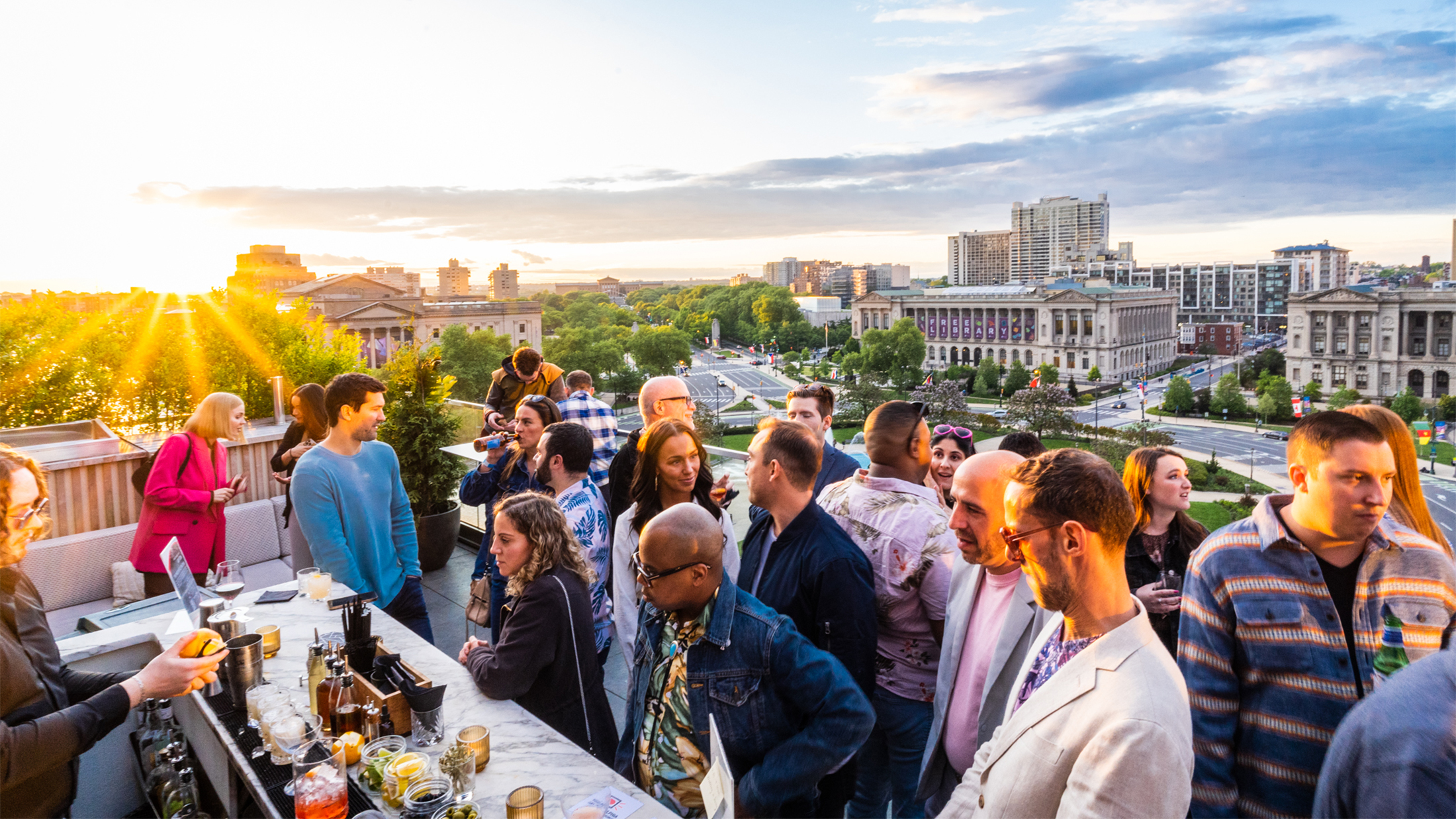 People order drinks at Assembly Rooftop Lounge with a view of the Parkway Central Library in the background as the sun sets.