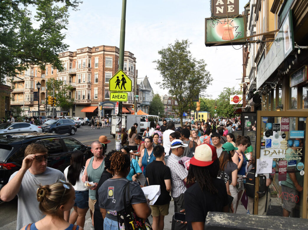 Crowds of people gather along Baltimore Avenue in Philadelphia during the Baltimore Avenue Dollar Stroll, lining the sidewalks outside neighborhood restaurants and shops on a busy summer evening.