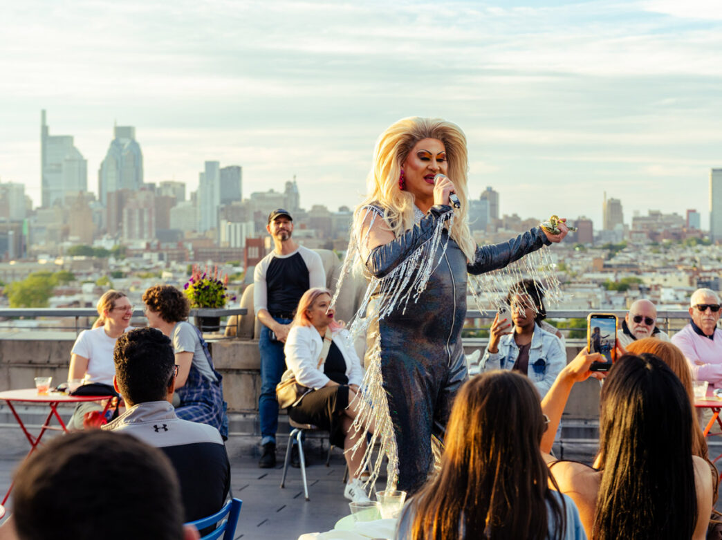A drag performer in a black outfit with sparkling fringe speaks into a microphone at Bok Bar with a view of the Philadelphia skyline in the background.