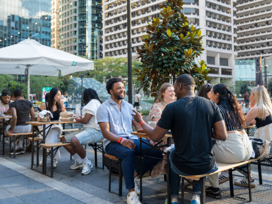 A group of people enjoy drinks and socialize at outdoor tables in Dilworth Park in downtown Philadelphia, with tall skyscrapers and trees visible in the background.