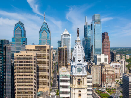 Philadelphia's Center City skyline, featuring the William Penn statue atop City Hall surrounded by modern glass skyscrapers beneath a bright blue sky.