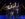 A line of dancers in black costumes perform onstage with arms raised with jazz hands, led by a dancers in a shorty black dress under bright theatrical lighting.
