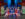 Performers dressed as festive toy soldiers in red, blue and gold costumes stand in a V formation with arms outstretched on a brightly colored holiday-themed stage featuring oversized gingerbread figures and Christmas trees.