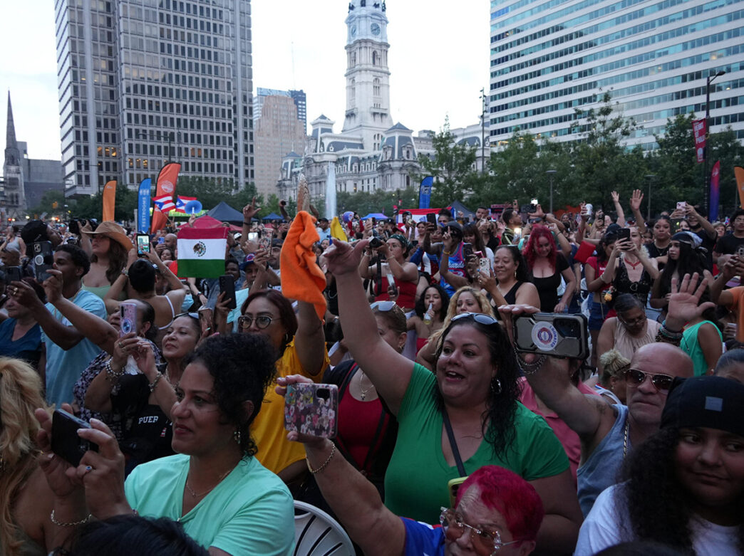 A large, diverse crowd cheers and records a live outdoor performance in front of Philadelphia's City Hall, with flags, raised hands and phones visible throughout the audience.