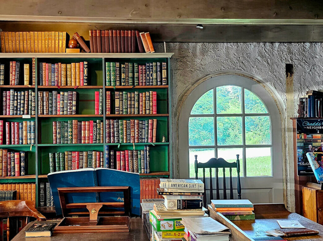 The interior of Baldwin's Book Barn, with leather-bound books on a green bookshelf and a pile of books and tabletop games stacked on a table in the foreground.