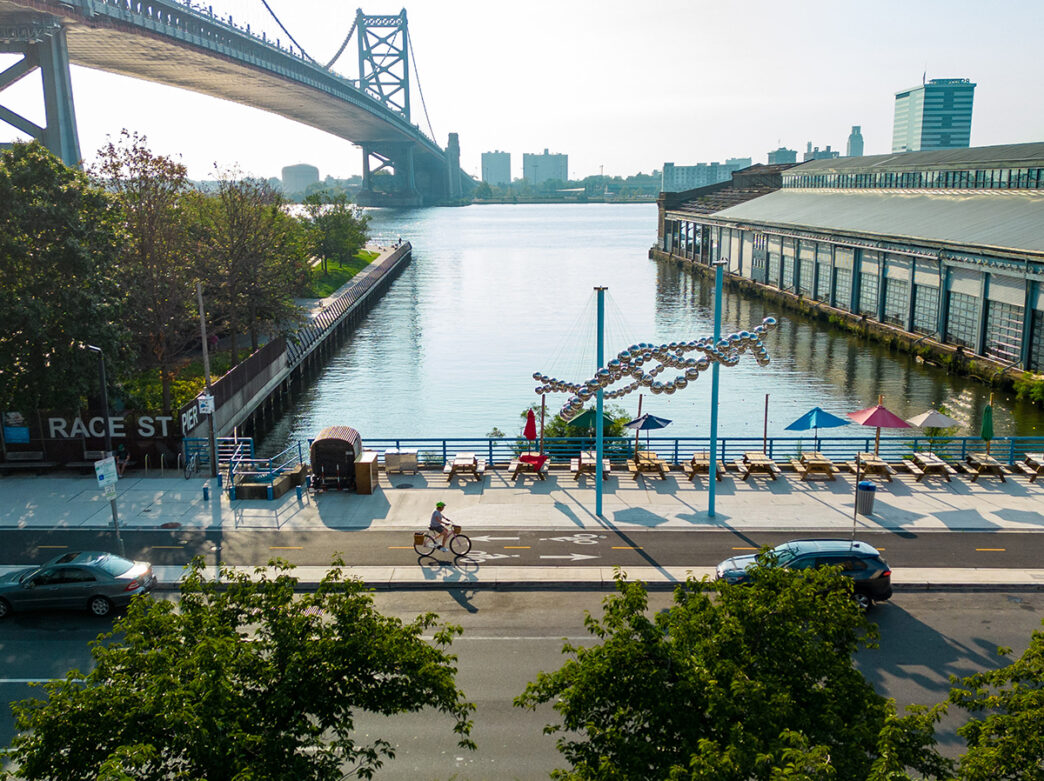 A biker wearing a helmet rides along the Delaware River Trail in front of the Race Street Pier, Cherry Street Pier and Benjamin Franklin Bridge.