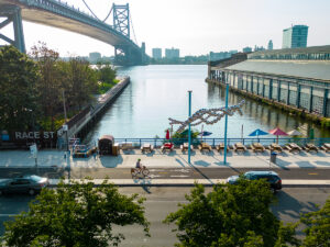A biker wearing a helmet rides along the Delaware River Trail in front of the Race Street Pier, Cherry Street Pier and Benjamin Franklin Bridge.