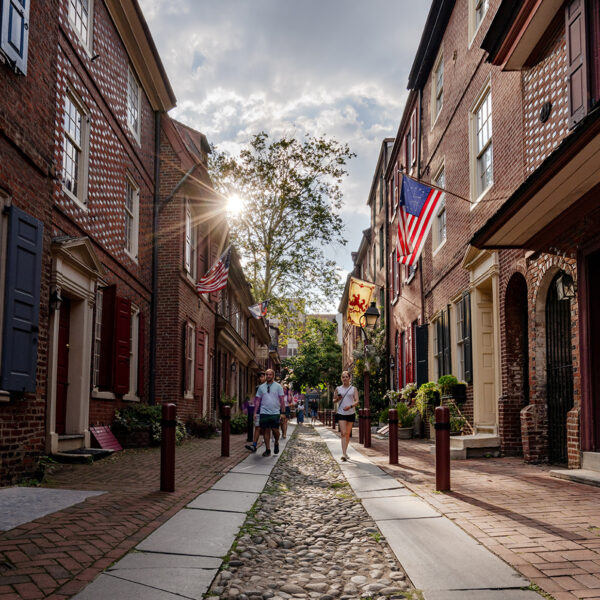 People walk along the cobblestone streets of Elfreth's Alley in Philadelphia with the sun rising up from behind a house.