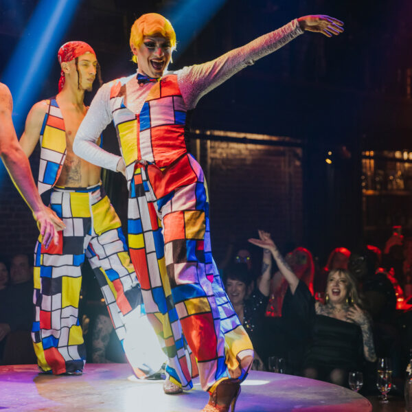 Performers in brightly colored geometric-patterned costumes dance on a circular stage at Fabrika in Philadelphia, illuminated by dramatic spotlights as audience members watch and cheer from surrounding tables.