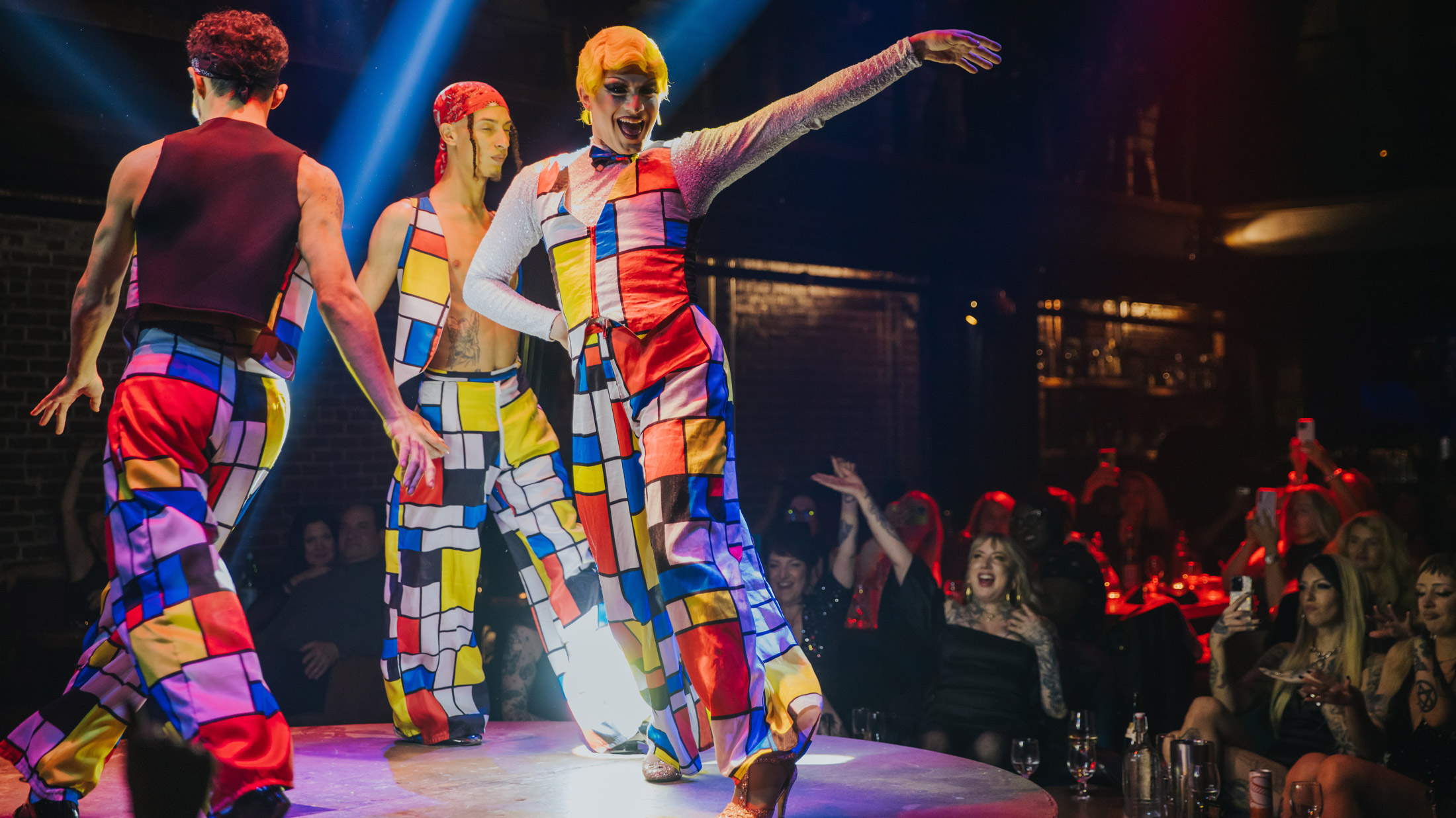 Performers in brightly colored geometric-patterned costumes dance on a circular stage at Fabrika in Philadelphia, illuminated by dramatic spotlights as audience members watch and cheer from surrounding tables.