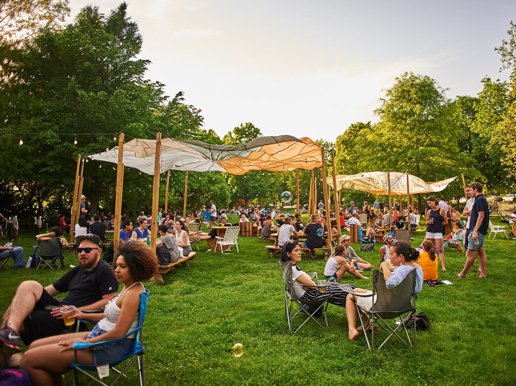 People relax in lawn chairs and picnic tables on a grassy lawn under fabric canopies at a pop-up beer garden in Fernhill Park.