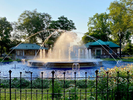 Water sprays from the circular fountain at Franklin Square as sunlight glows through the mist, with flowers, trees and park buildings surrounding the fountain.