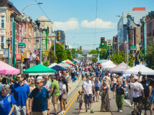 Crowds of people stroll along a closed-off Girard Avenue during the Girard Avenue Street Fest in Philadelphia's Brewerytown neighborhood, lined with colorful vendor tents and historic rowhouses on a sunny day.