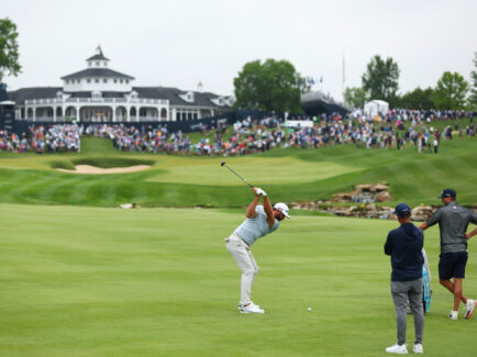 A golf swings at a golf ball on the fairway. Two caddies stand with the golfers bag. A crowd of people watching can be seen in the far distance.