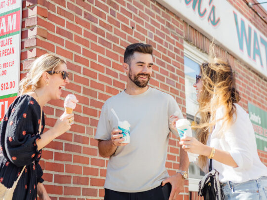 Three people stand in a group outside John's Water Ice laughing and eating water ice and ice cream