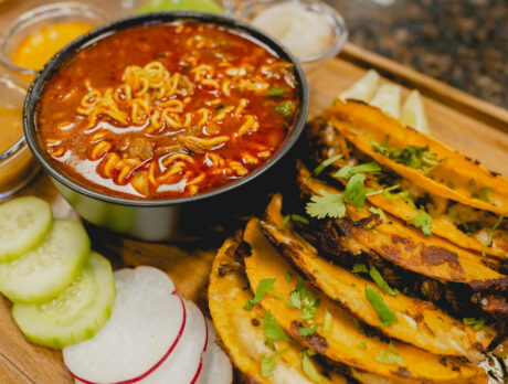 Tacos sit on a table next to fresh vegetables and a birria ramen soup at La Catrachita Taqueria
