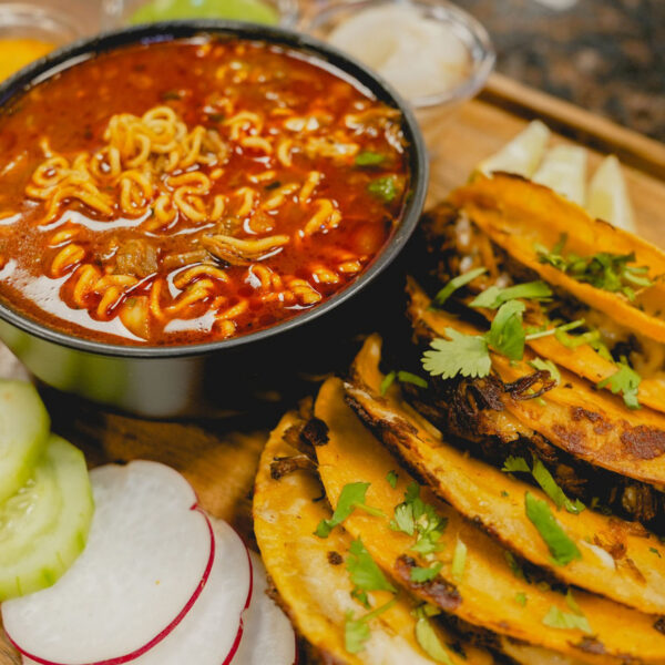 Tacos sit on a table next to fresh vegetables and a birria ramen soup at La Catrachita Taqueria