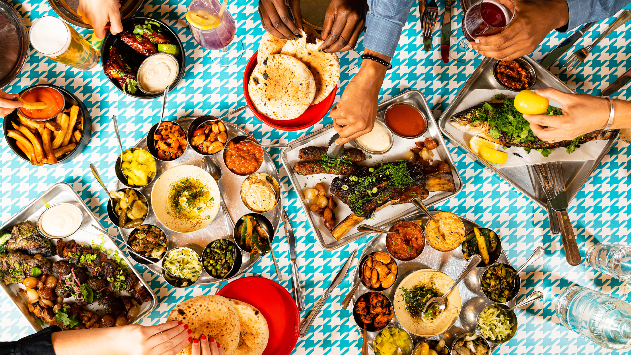 A spread of Laser Wolf menu items set against a teal and white houndstooth pattern. The dishes include fries with ketchup, grilled meats, whole fish with herbs and lemon, pita, an assortment of hummus and other dips. Diners' hands can be seen reaching for food.