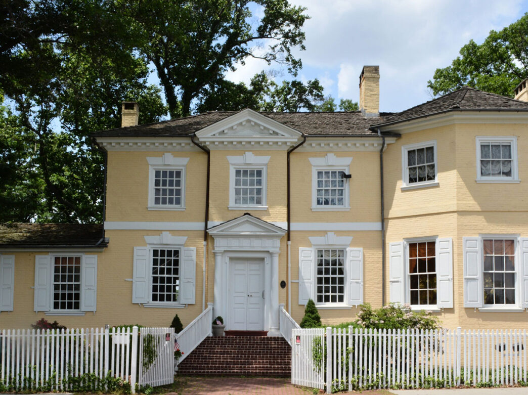 A yellow historic mansion with white shutters and a white doorway, framed by white picket fence and shaded by tall trees.