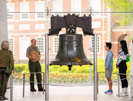 A National Park Service ranger leads a tour inside the Liberty Bell Center as visitors of different ages stand around the Liberty Bell listening to the presentation. The historic cracked bell is centered in the gallery, with Independence Hall visible through the floor-to-ceiling windows behind it.
