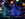 Brightly lit trees glow in purple, blue and green, framed by strands of colorful bulbs, glow against the dark sky.