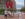 Visitors take photos at the iconic red LOVE sculpture in Philadelphia's Love Park. The large letters are stacked with blue and purple elements visible through the "O" and "E." Historic brick and modern high-rise buildings surround the plaza, with green landscaping, a fountain, and red chairs visible in the background on a partly cloudy day.