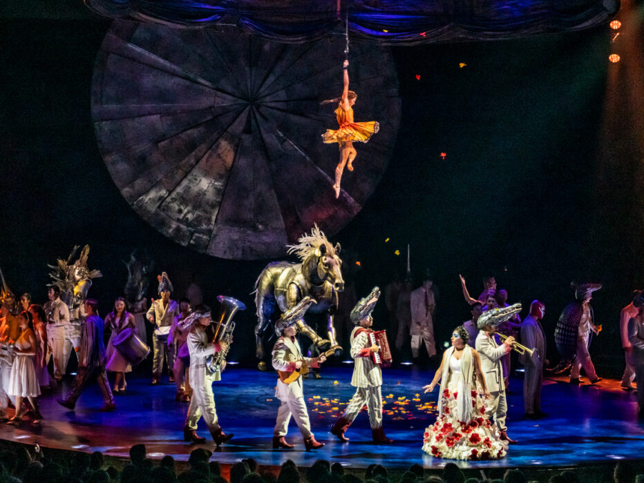 Cirque du Soleil's LUZIA performance featuring a diverse cast of acrobats and performers in colorful costumes on stage, with a large golden horse prop, an aerial performer suspended from above against a geometric backdrop, and dramatic blue and orange stage lighting.