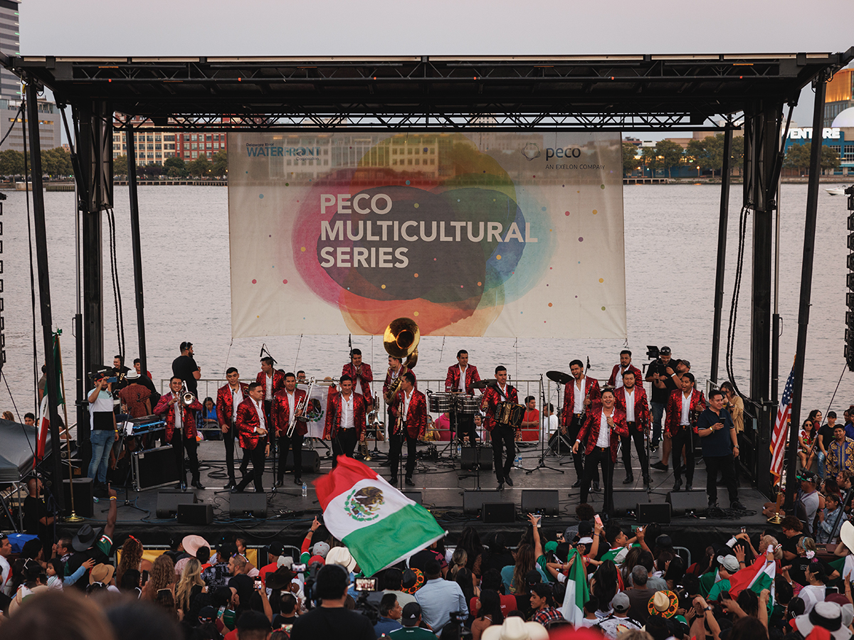 A band plays on a stage for Mexican Independence Day Festival in front of the Delaware River while festivalgoers dance and listen to music.