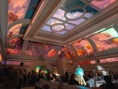 A group of visitors gather in the ornate Hall at Ministry of Awe in Philadelphia, gazing upward at an immersive ceiling installation. The historic beamed ceiling displays vibrant projected imagery in oranges, pinks, purples, and blues, creating an awe-inspiring visual experience. Architectural details include decorative molding and arched doorways, while ambient lighting enhances the contemplative atmosphere.