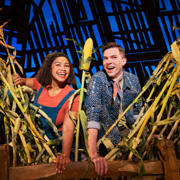 Two performers smile and lean over a wooden fence surrounded by tall cornstalks on a barn-themed stage set from the musical Shucked.