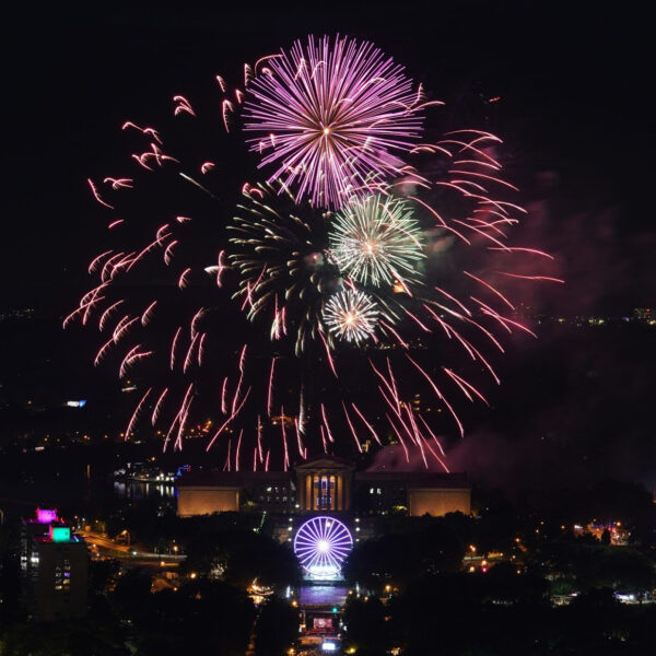 A glowing fireworks display explodes over the Philadelphia Museum of Art and Benjamin Franklin Parkway in a night sky.