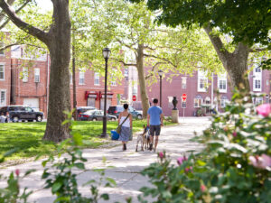 A couple walks their dogs along a sunny, tree-lined path through a park in Philadelphia's Pennsport neighborhood. Brick row homes and cars parked on the road are in the background.
