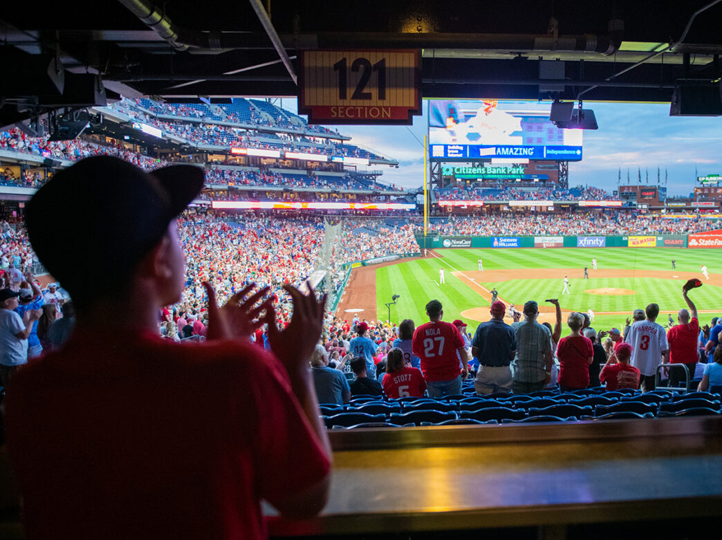 A young fan watching the game from a shaded section at Citizens Bank Park claps enthusiastically, joining the lively crowd as they cheer on the Phillies.