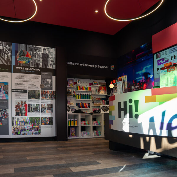 An interior view of the Philly Pride Visitor Center showing colorful displays on LGBTQ+ history, a bright welcome desk and Pride merchandise displays beneath modern circular light fixtures.