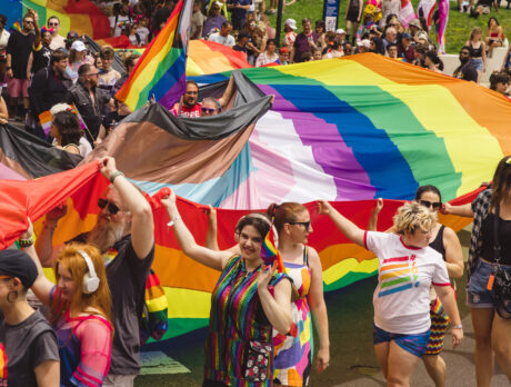 Participants march through Philadelphia’s Pride celebration carrying large rainbow and LGBTQ+ pride flags, surrounded by a dense crowd of spectators and marchers in colorful clothing and accessories.