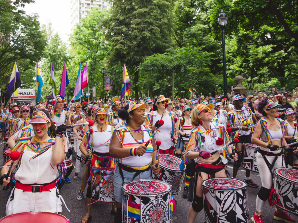 A colorful drumline performs during the Philadelphia Pride March as musicians in rainbow-trimmed outfits march down a tree-lined street. Crowds gather along the route waving pride flags and cheering during the outdoor celebration.