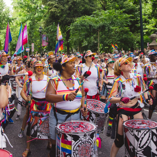 A colorful drumline performs during the Philadelphia Pride March as musicians in rainbow-trimmed outfits march down a tree-lined street. Crowds gather along the route waving pride flags and cheering during the outdoor celebration.