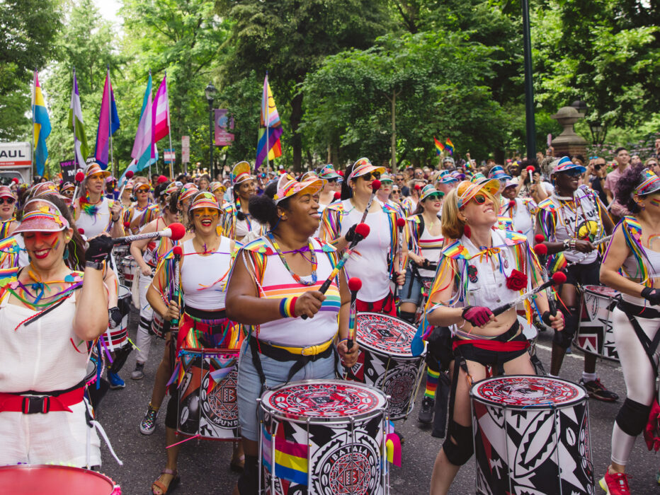A colorful drumline performs during the Philadelphia Pride March as musicians in rainbow-trimmed outfits march down a tree-lined street. Crowds gather along the route waving pride flags and cheering during the outdoor celebration.