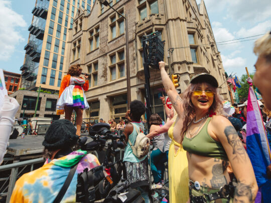 People celebrate at a Philadelphia Pride street festival as a performer sings on an outdoor stage. In the foreground, attendees wave rainbow and bisexual pride flags and dance in front of historic Center City buildings on a sunny day.