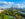 A wide aerial view shows Philadelphia's skyline beyond a tree-filled park and the Schuylkill River, with blue skies and fluffy clouds overhead.