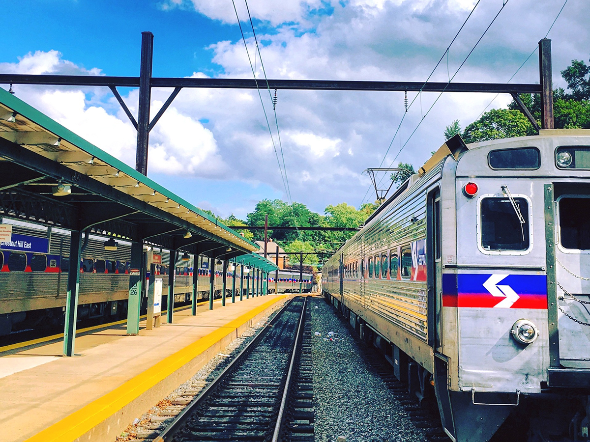 Two silver SEPTA Regional Rail trains wait at the Chestnut Hill East Station in Philadelphia, next to the bright yellow platform edges and green canopy roofs of the platform.