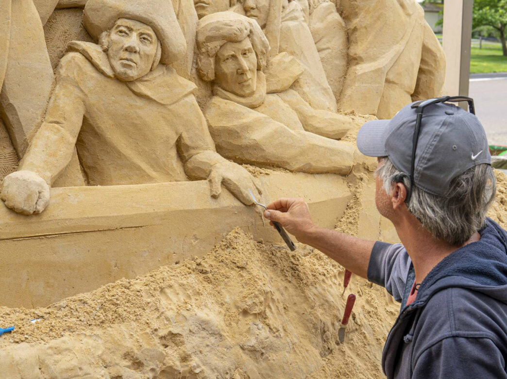 An artist carefully carves details into a large sand sculpture at Peddler’s Village’s “Sights in Sand: An American Road Trip” exhibit, depicting historical figures emerging from the sculpted scene.