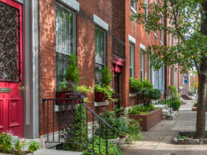 A charming tree-lined block of red brick rowhouses in Philadelphia's Spring Garden neighborhood, featuring lush window boxes, ornate ironwork railings, and a bold red front door.