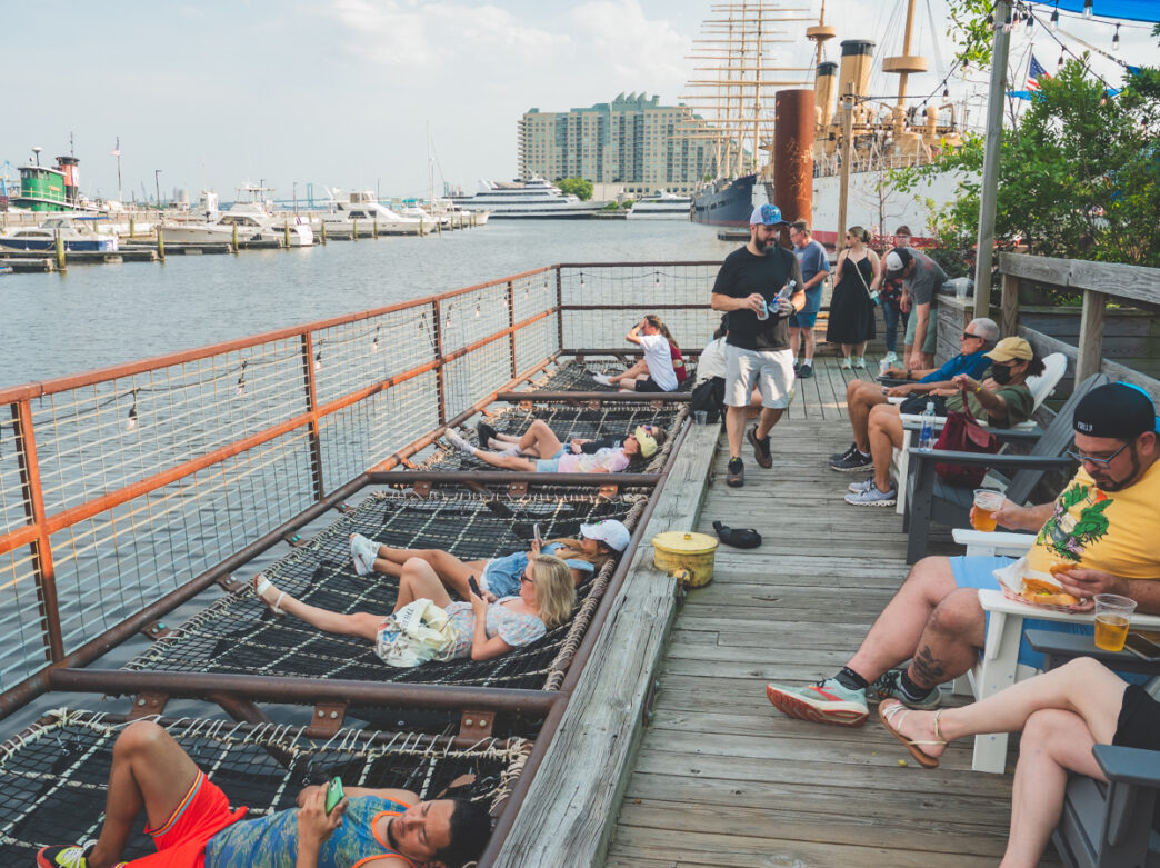 Visitors relax in a netted lounge area along floating barges at Spruce Street Harbor Park in Philadelphia, overlooking the Delaware River on a sunny day.